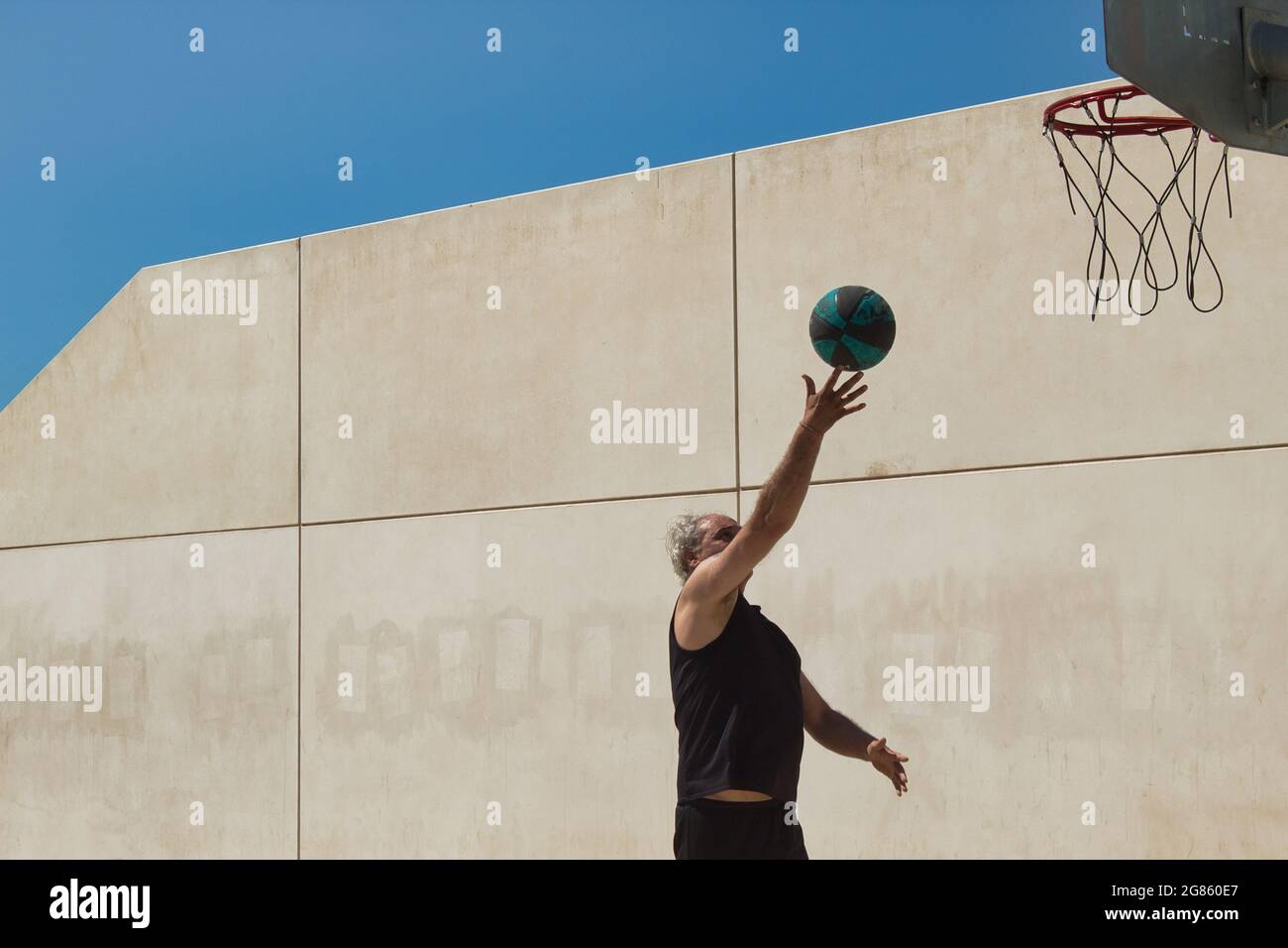 homme mature jouant au basket-ball seul sur un terrain de rue avec panier en métal et mur en arrière-plan Banque D'Images