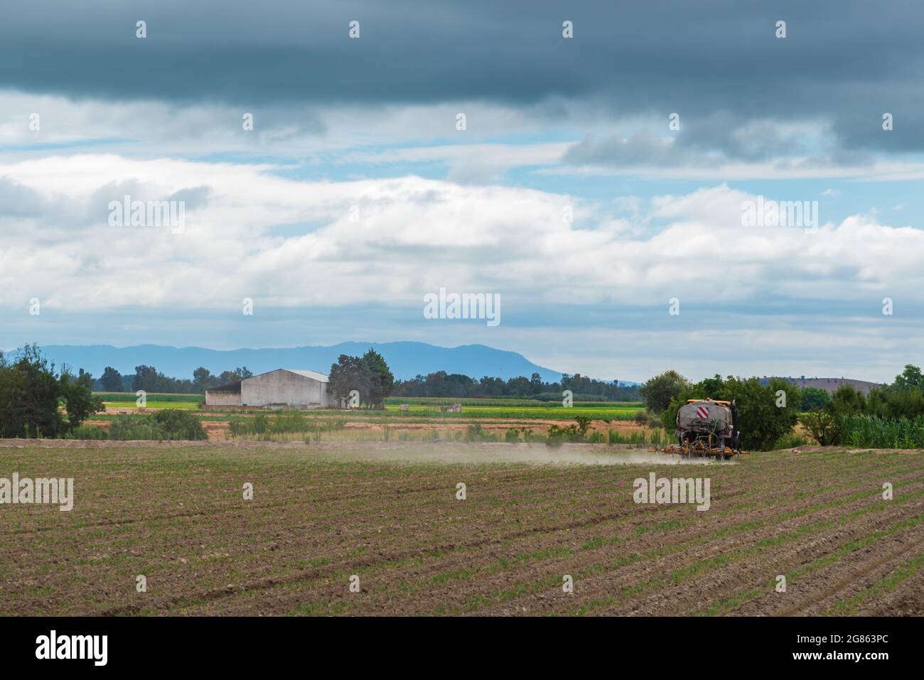 Tracteur agricole travaillant dans le champ, avec un paysage vert, un entrepôt industriel agricole, des montagnes et le ciel nuageux. Banque D'Images