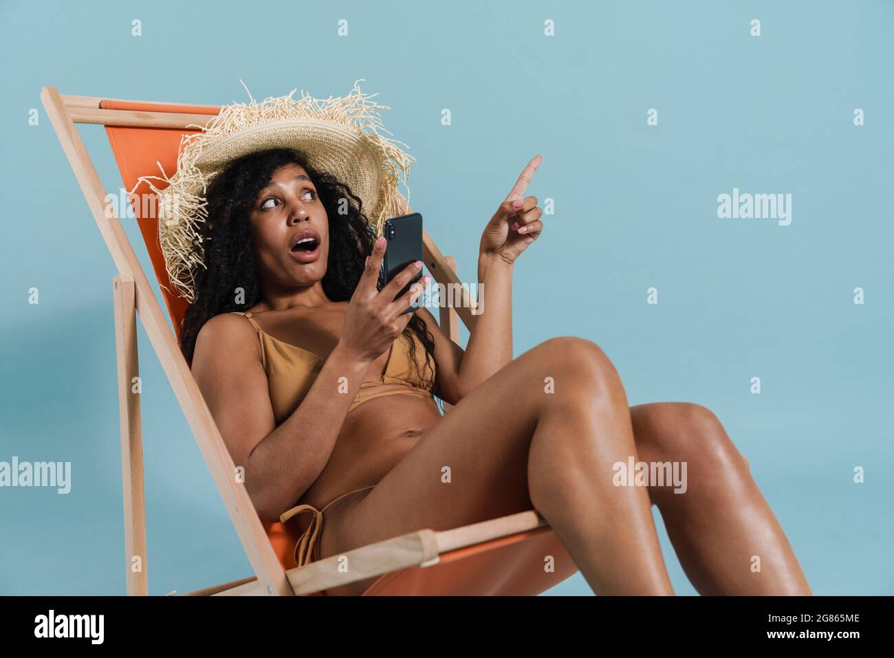 Noire choquée femme utilisant le téléphone portable tout en étant assis dans une chaise longue isolée sur fond bleu Banque D'Images