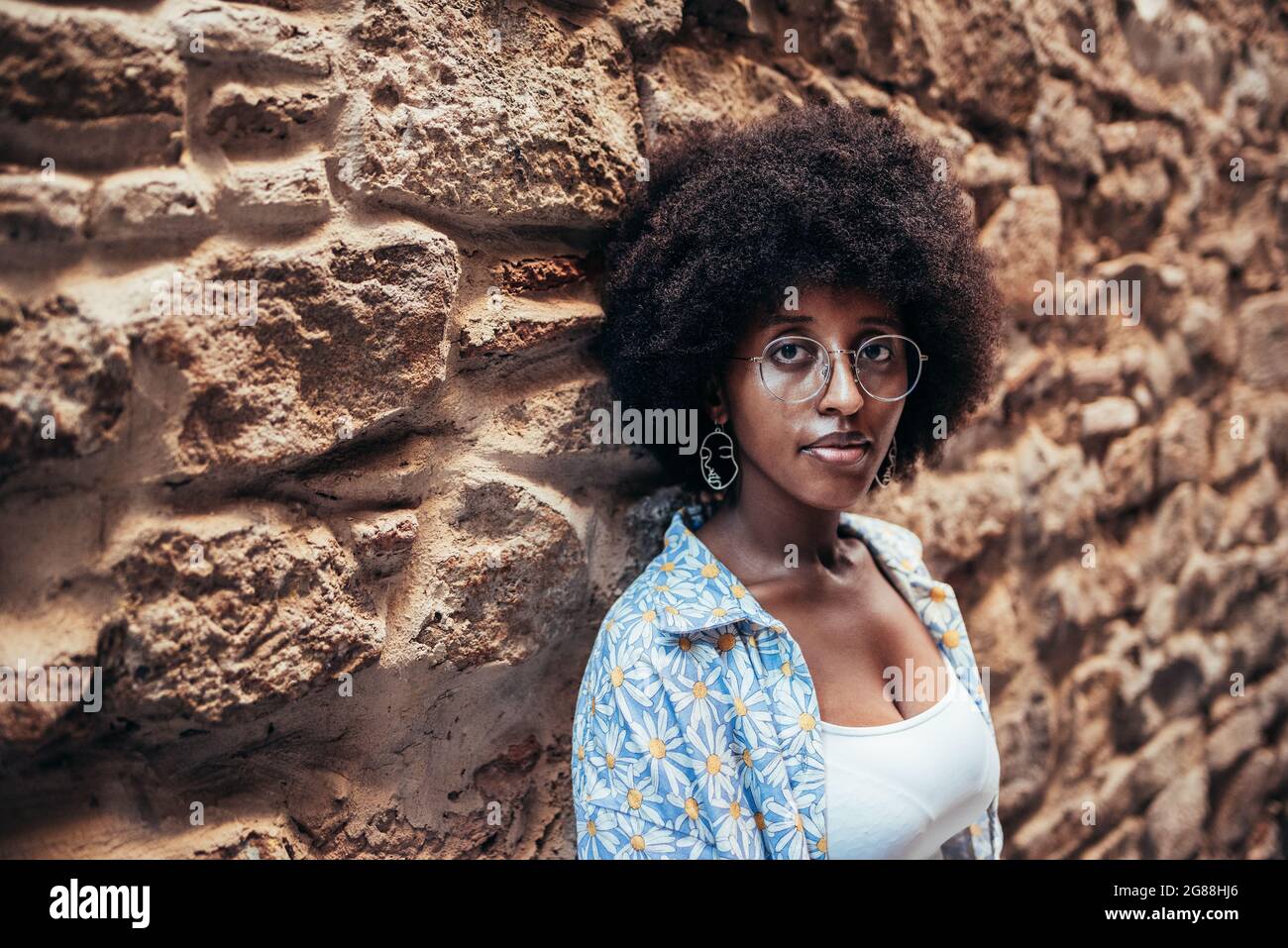 femme afro avec des lunettes et des vêtements décontractés regardant l'appareil photo. Elle est penchée sur un mur de pierre Banque D'Images