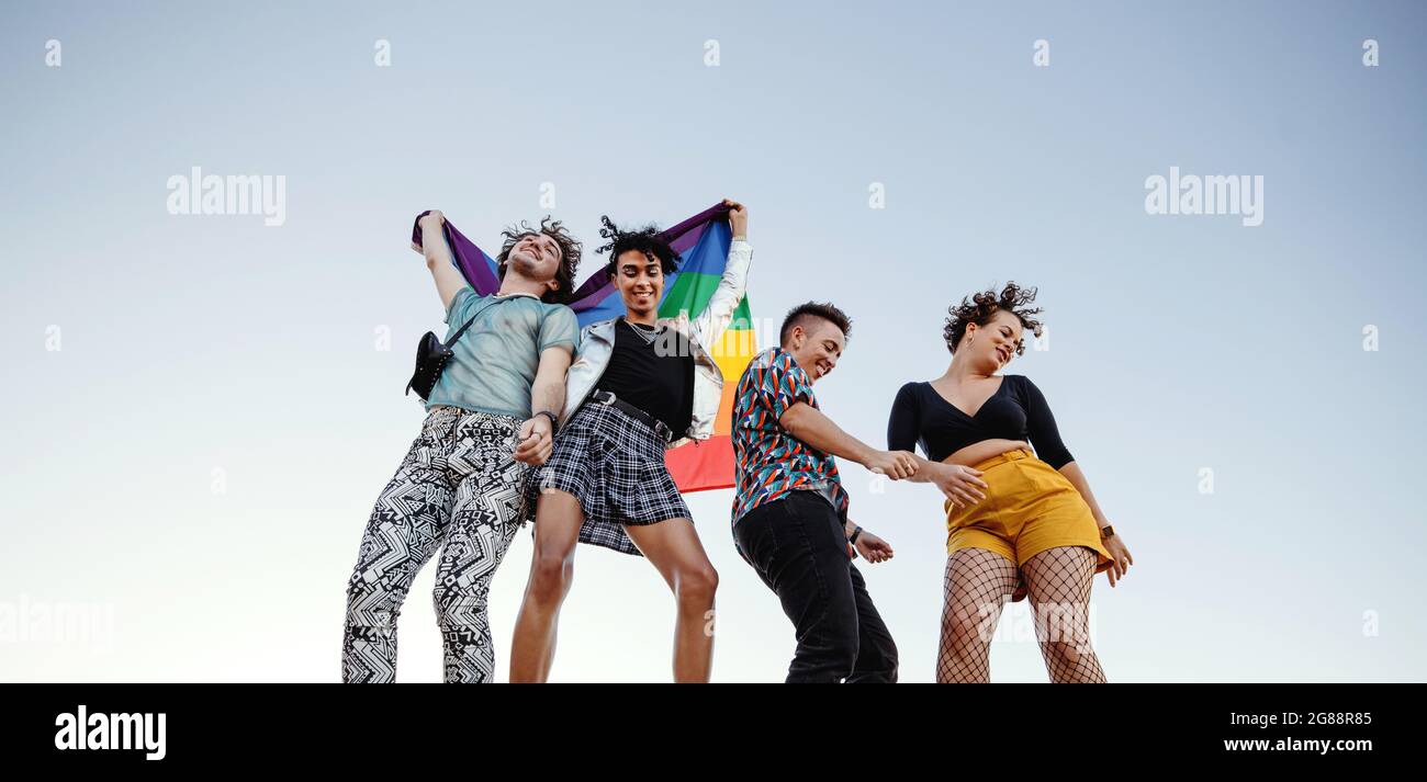 Groupe joyeux d'individus queer célébrant la fierté gay. Quatre membres de la communauté LGBTQ+ dansent avec joie tout en levant le drapeau de la fierté de l'arc-en-ciel. Banque D'Images