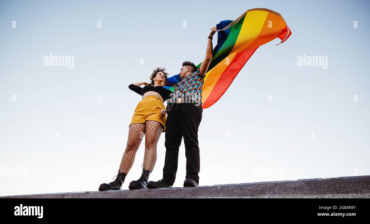 Couple LGBTQ élevant le drapeau arc-en-ciel à l'extérieur. Queer jeune homme et femme embrassant leur identité LGBTQ+. Jeune couple non conforme debout W Banque D'Images