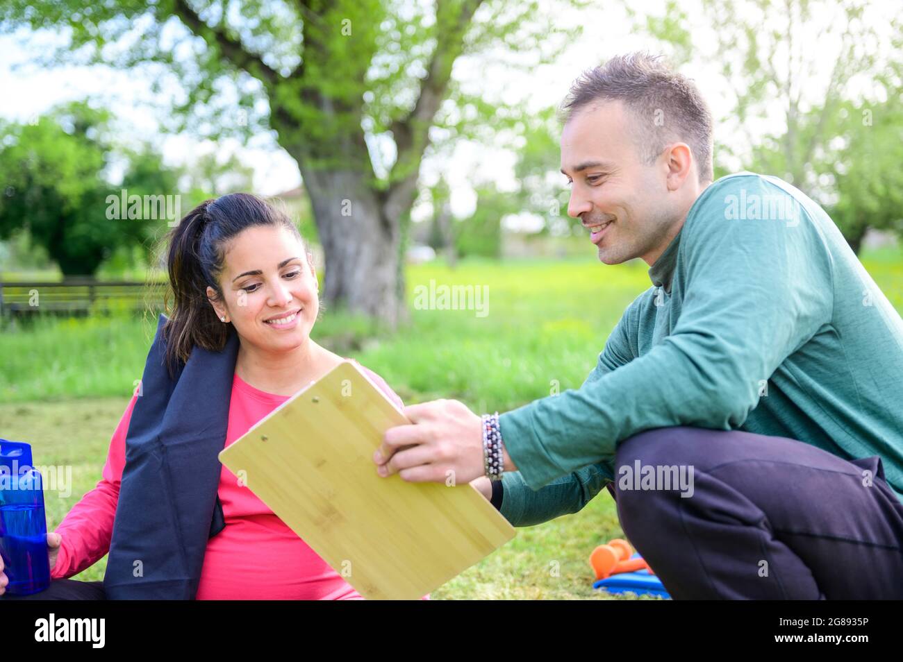 Entraîneur personnel et une jeune femme enceinte parlant le programme d'entraînement ensemble et souriant dans un parc extérieur - concept de forme physique et de mode de vie sain, Banque D'Images
