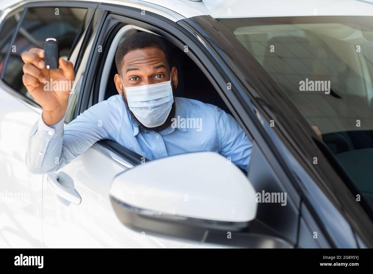 Homme africain heureux dans le masque médical assis en voiture et montrant la clé Banque D'Images