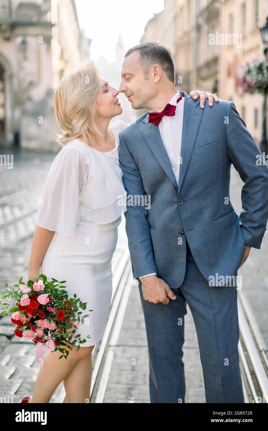 Gros plan portrait d'un couple aimant, d'un homme beau de 50 ans en costume  et noeud papillon, debout avec sa jolie femme en robe blanche avec bouquet  de fleurs, sur le fond