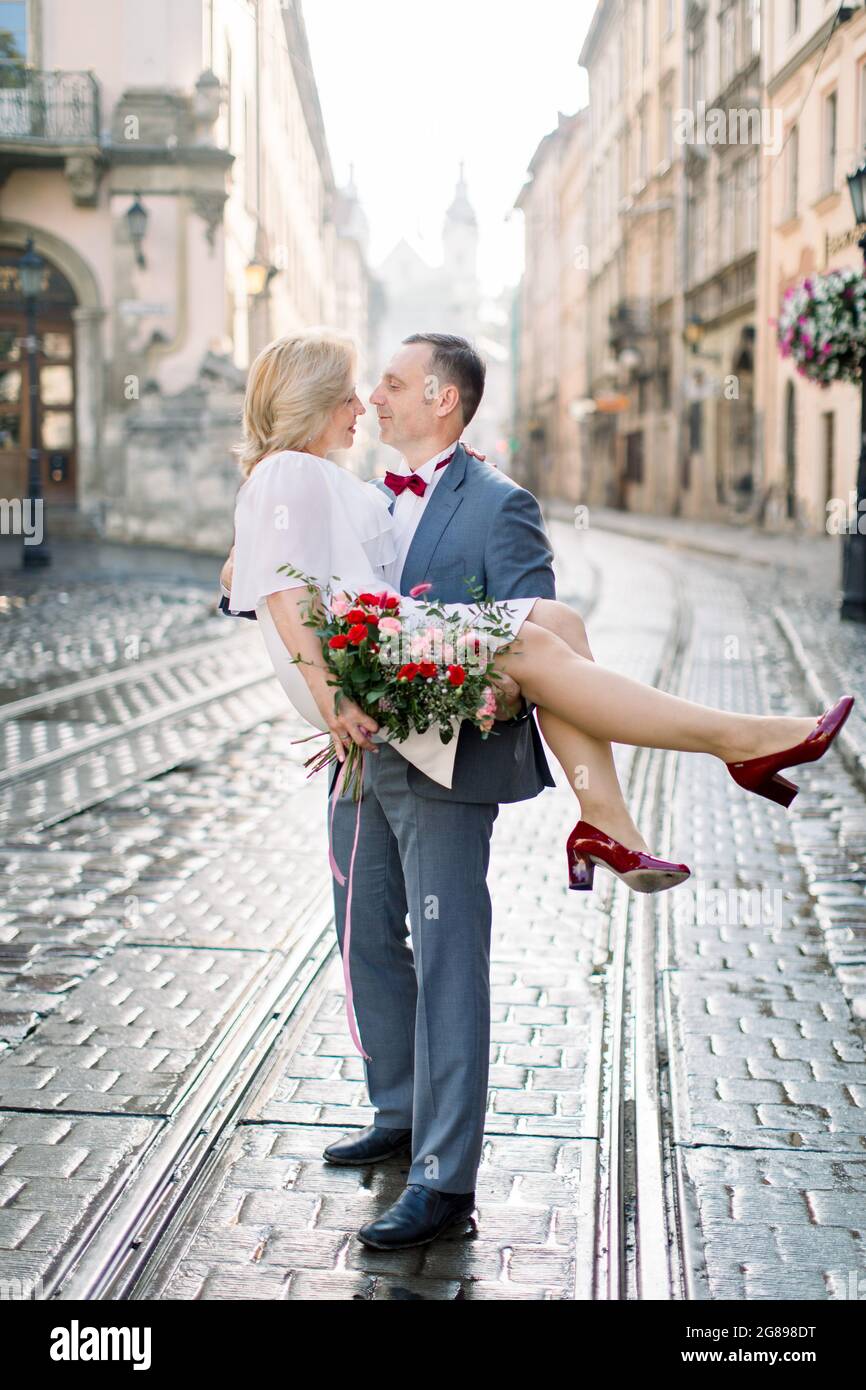 Portrait complet d'un beau couple mature dans la vieille ville antique. Beau homme en costume tient la charmante femme blonde sur les mains. Anniversaire de mariage Banque D'Images