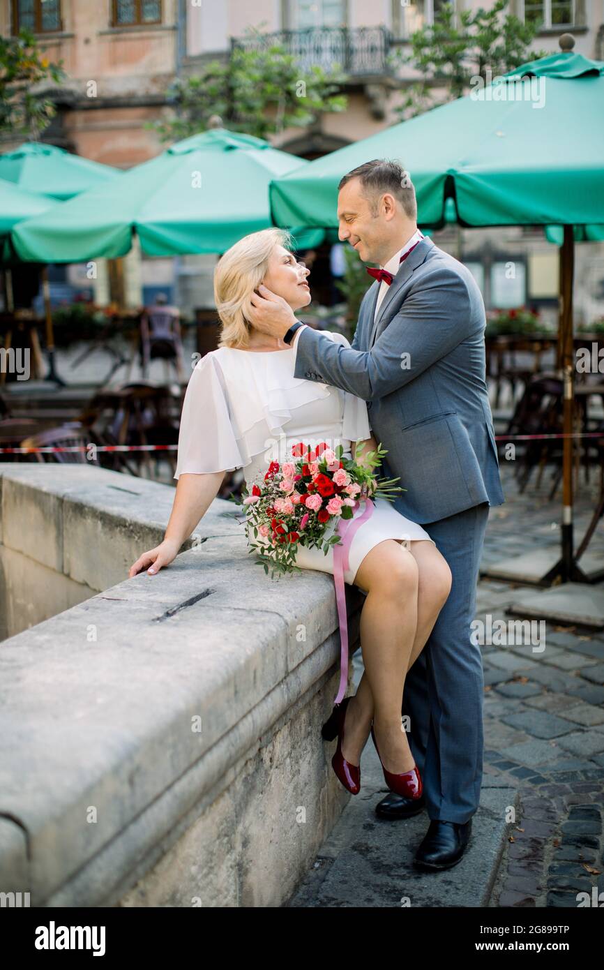 Beau couple d'âge mûr romantique profitant de moments tendres entre eux.  Femme en robe blanche est assise sur la vieille ville en pierre fontaine  avec un bouquet de fleurs, l'homme touche son