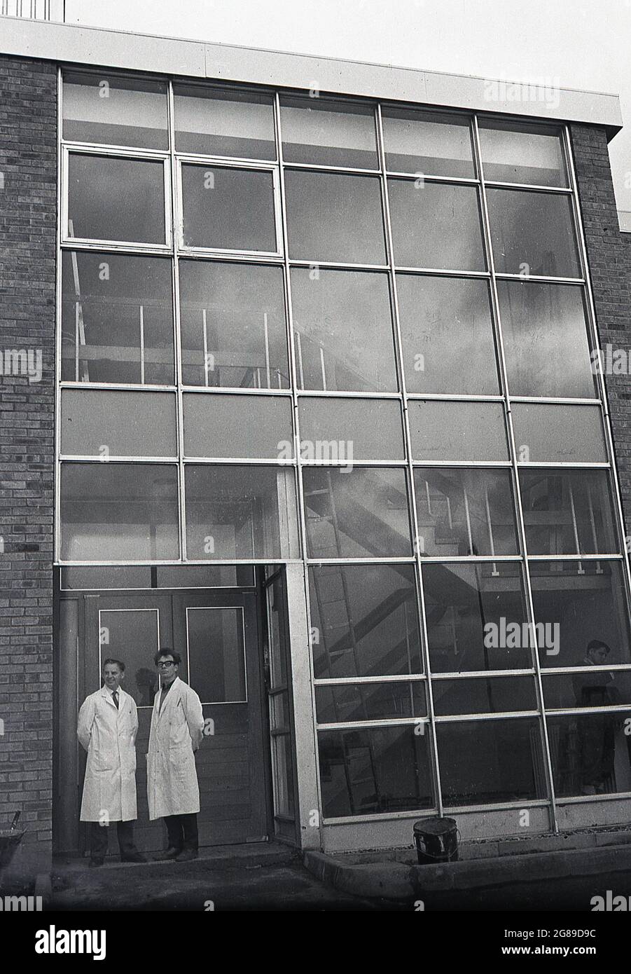 Années 1960, historique, deux chercheurs en sciences de la recherche de sexe masculin blancooté se tenant à l'extérieur d'une entrée d'un escalier ou d'un escalier extérieur mais clos, dans un immeuble de bureaux moderne, typique de l'époque, avec son panneau extérieur de fenêtre, Angleterre, Royaume-Uni. Ces grandes fenêtres à panneaux ont été fabriquées à l'extérieur, ont été produites en masse et ont donc rendu les coûts de construction moins chers, réduisant le besoin de main-d'œuvre qualifiée comme des briques à utiliser. Le problème des panneaux de verre était qu'au fil du temps ils ne durent pas ou vieillissaient bien, après avoir été produits à bon marché, ils ont subi des ruptures et des fuites et donc les coûts d'entretien étaient élevés. Banque D'Images