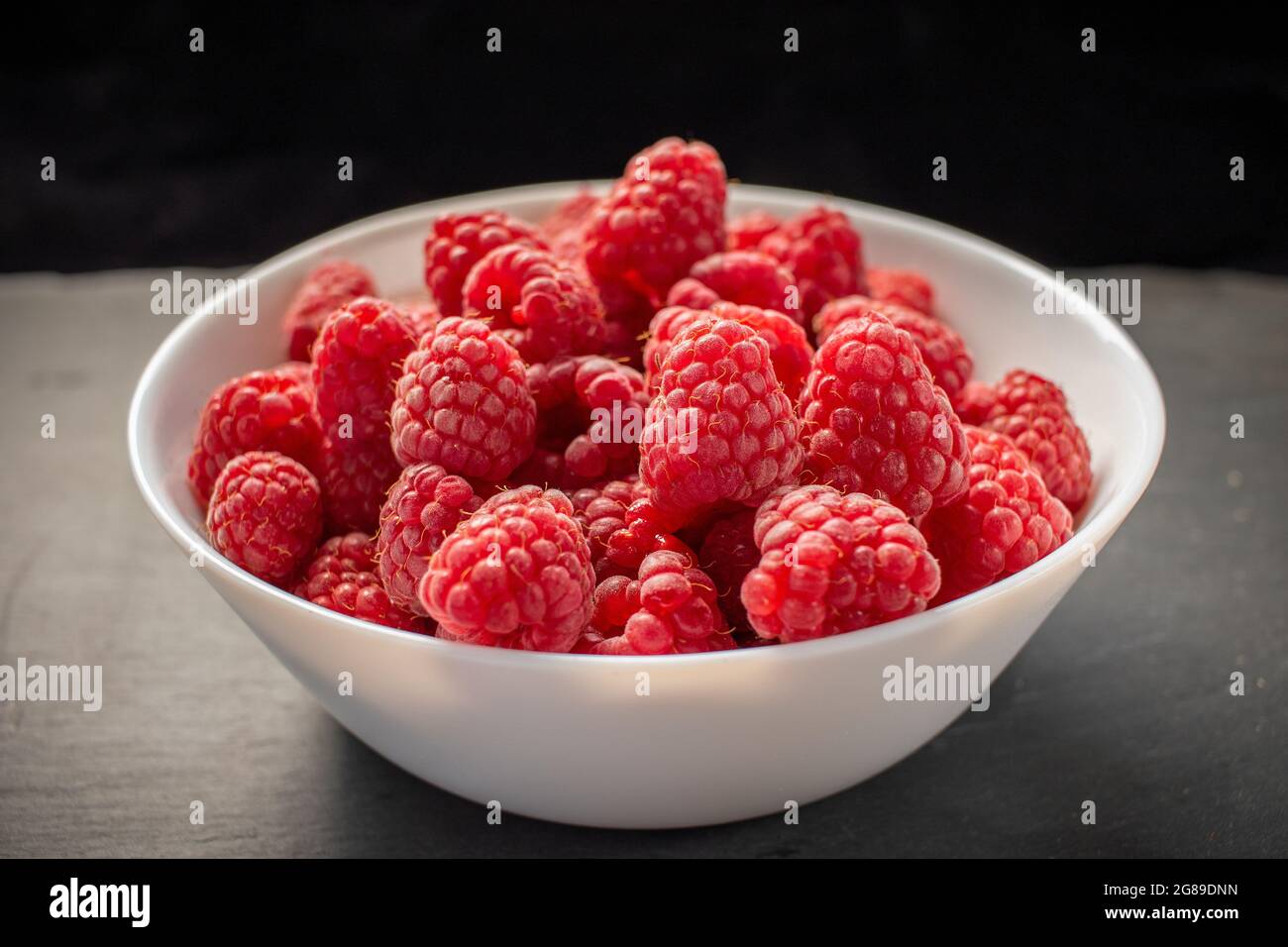 Framboises dans un bol blanc sur fond de pierre noire Banque D'Images