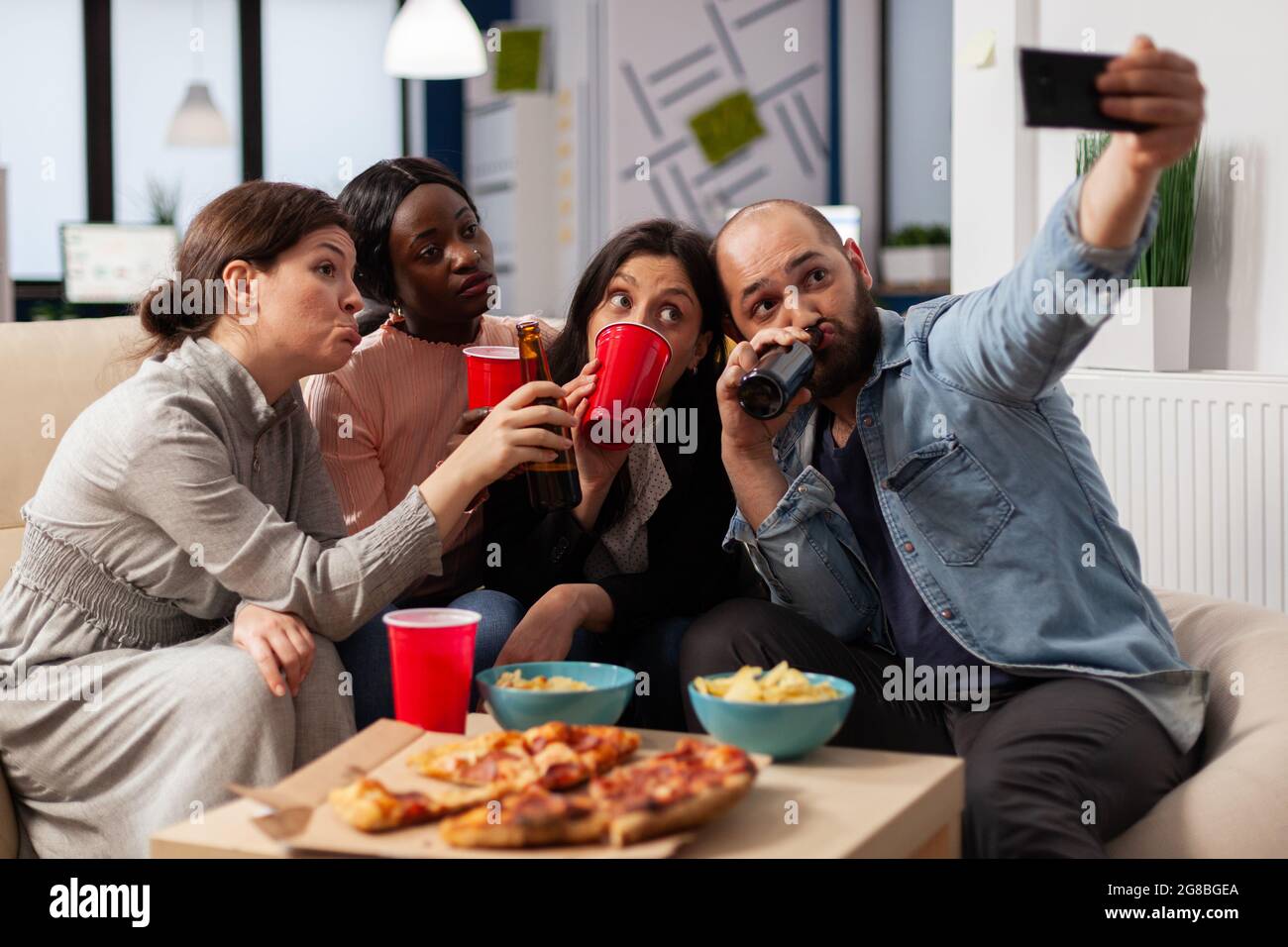 Une équipe d'amis multi-ethniques se fait des souvenirs après une fête de travail en prenant des photos sur un smartphone. Collègues fêtant dans un espace de bureau avec en-cas pizzas frites tasses de boissons sur table Banque D'Images