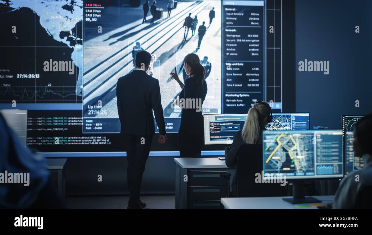 Des fonctionnaires gouvernementaux masculins et féminins discutent de questions de travail à côté d'un ordinateur et d'un grand écran numérique avec des images de surveillance par satellite avec le visage Banque D'Images
