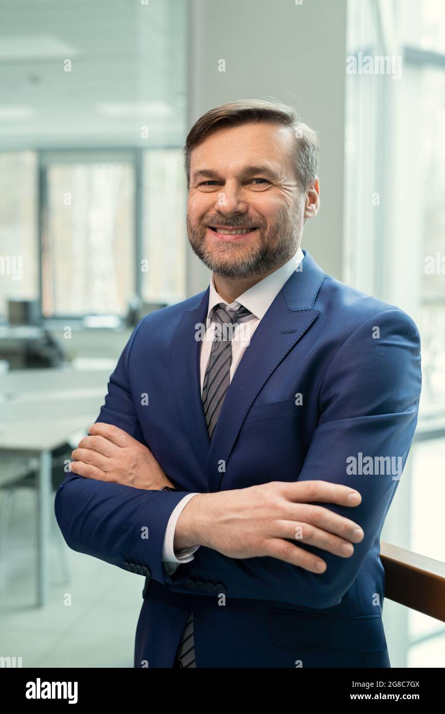 Portrait d'un homme d'affaires mature et réussi en costume élégant souriant à l'appareil photo tout en se tenant au bureau Banque D'Images