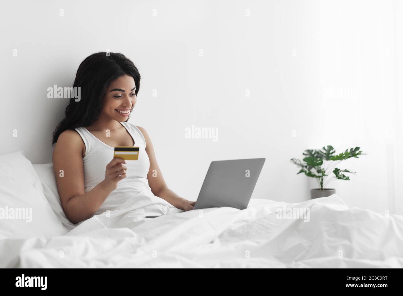 Concept de commerce électronique. Joyeux jeune afro-américain assis au lit, payant avec carte de crédit par ordinateur portable, espace libre Banque D'Images