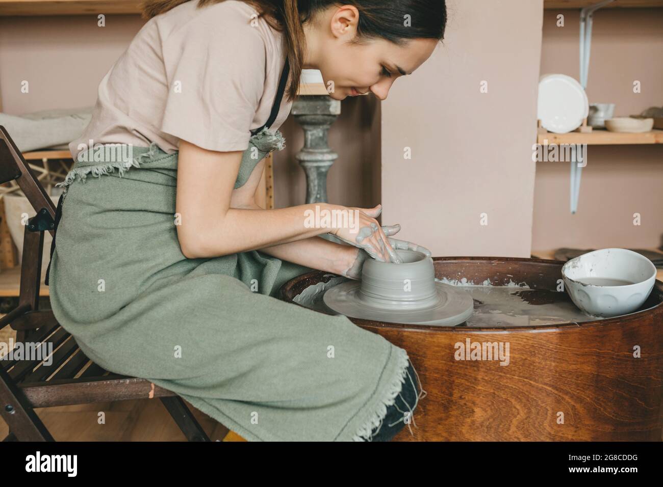 La jeune femme poterie travaille en studio d'argile sur la roue de poterie contre des étagères avec vases et pots. Concept de personnes créatives, petite entreprise. Banque D'Images