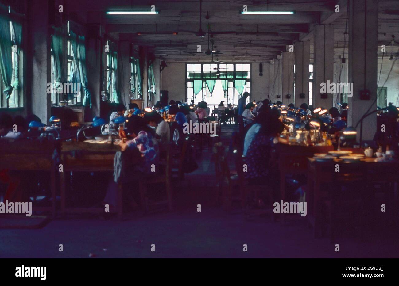 Chine 1983 une usine de peinture en porcelaine où des dessins exquis sont appliqués laborieusement par des femmes travaillant sous des lumières dans une pièce faiblement éclairée. Banque D'Images