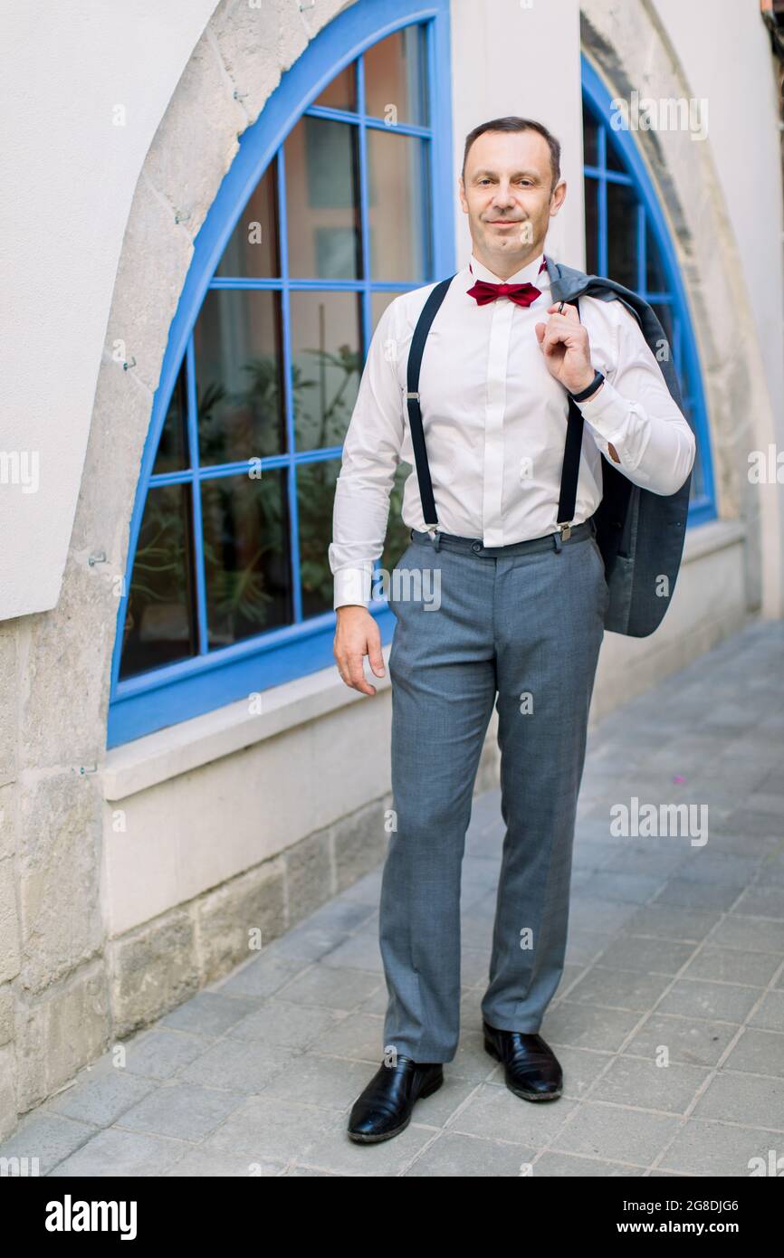 Beau caucasien mature homme marié dans une chemise blanche avec un noeud  rouge et dans les bretelles tient une veste, posant devant un bâtiment d' époque avec grande fenêtre bleue dehors dans la