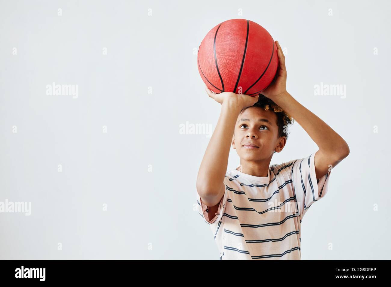 Portrait minimal d'un adolescent de course mixte qui jette un ballon de basket-ball tout en se tenant sur fond blanc, espace de copie Banque D'Images