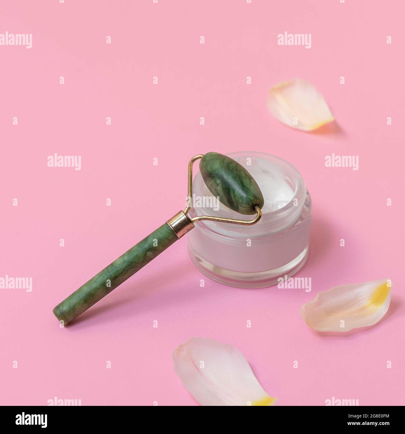 Massage du visage à roulettes Jade avec crème pour le visage sur rose pastel arrière-plan avec pétales blancs Banque D'Images