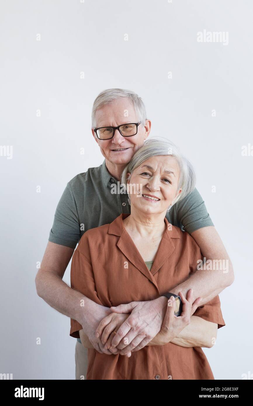 Portrait vertical d'un couple de personnes âgées aimant qui s'embrasse et regarde l'appareil photo tout en se tenant sur un arrière-plan blanc Banque D'Images