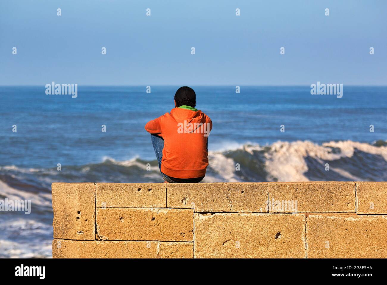 Jeune homme assis sur le mur de la ville, vue sur la mer, Essaouira, côte Atlantique, Marrakech-Safi, Maroc Banque D'Images