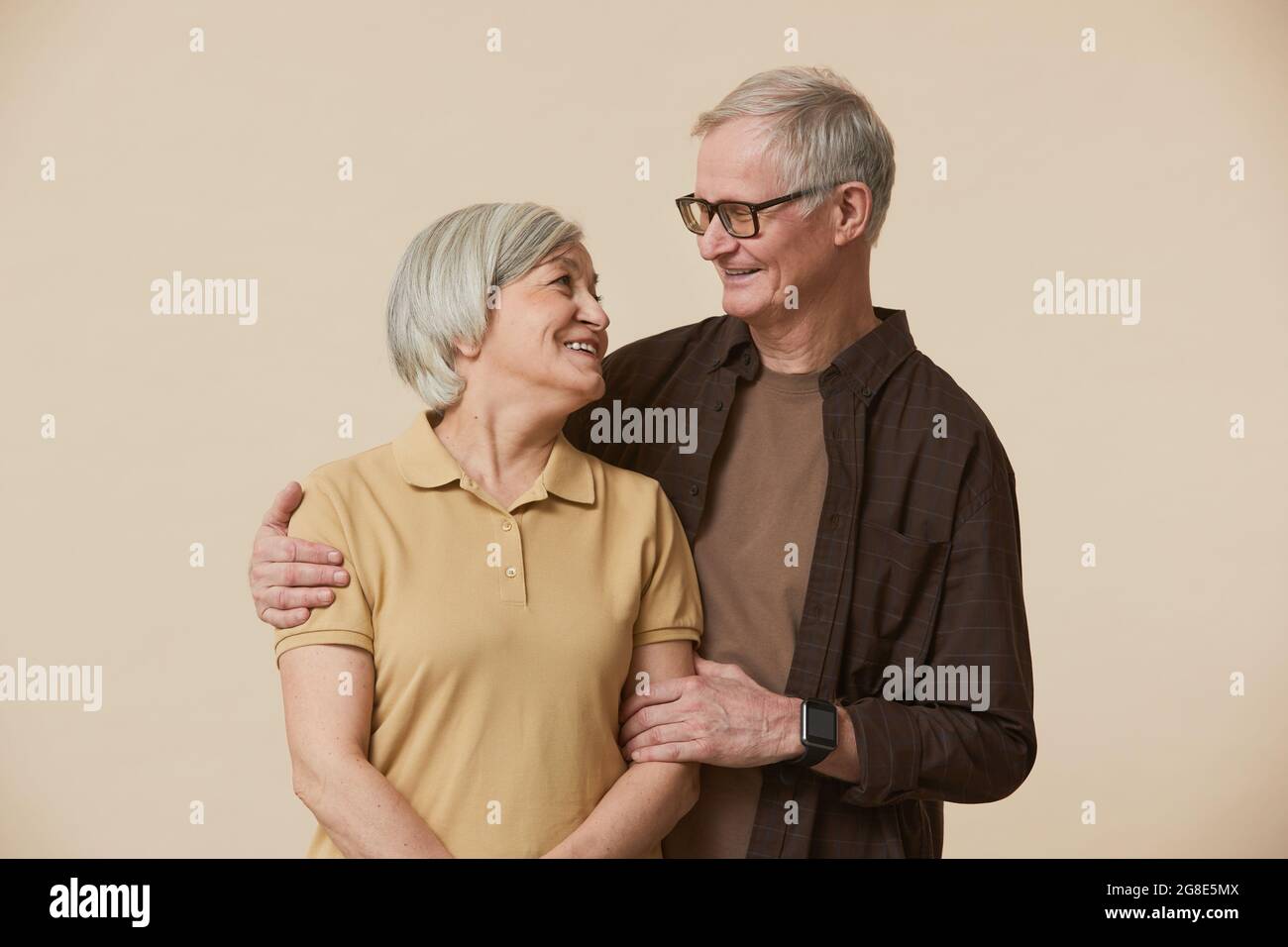 Portrait minimal de couple senior insouciant se tenant et regardant l'un l'autre tout en se tenant contre un arrière-plan beige Banque D'Images