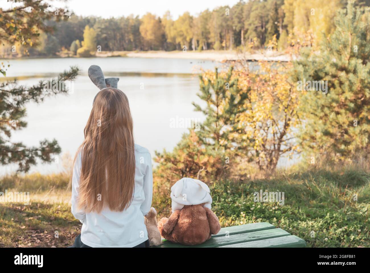 Une fille aux oreilles de lapin et à l'ours en peluche est assise sur un banc au lac Banque D'Images