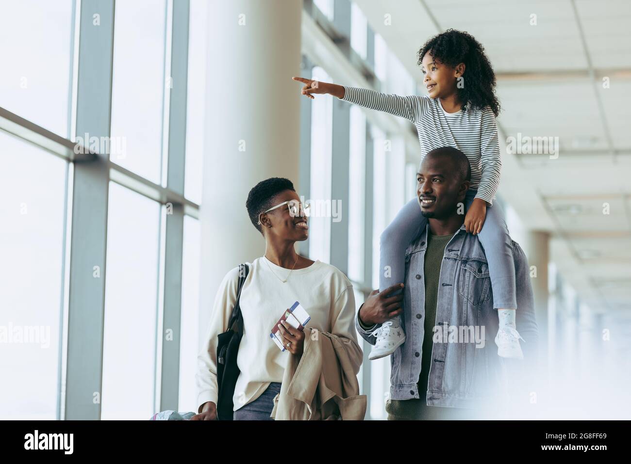 Bonne famille africaine en vacances à l'aéroport. Couple marchant à l'aéroport avec la fille assise sur l'épaule et pointant vers l'extérieur. Banque D'Images