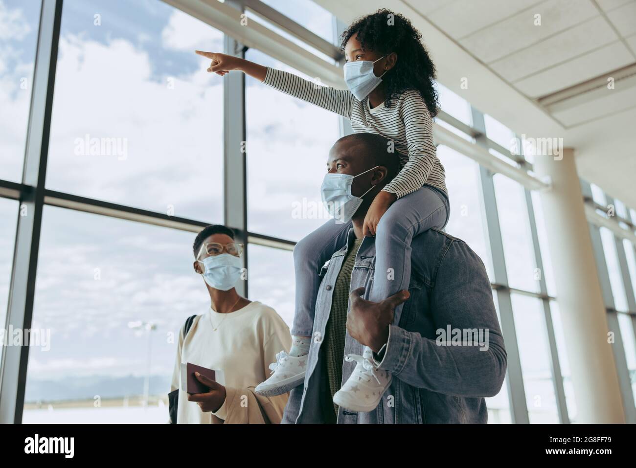 Père avec fille sur l'épaule et pointant des avions tout en marchant avec la mère à l'aéroport. La famille africaine à l'aéroport en pandémie. Banque D'Images