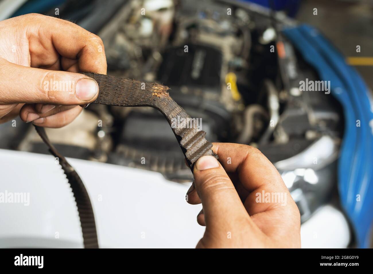 Un mécanicien d'automobiles montre une courroie de distribution déchirée avec des dents usées sur le fond d'un gros plan de capot de voiture ouvert Banque D'Images