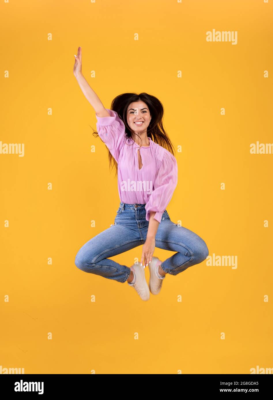 Joyeuse femme arménienne sautant et touchant les jambes avec la main, fond jaune de studio, pleine photo Banque D'Images