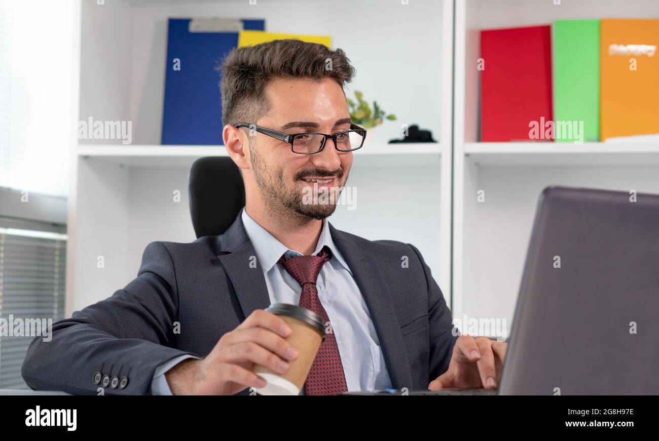 Portrait d'un homme d'affaires heureux dans des vêtements formels travaillant avec un ordinateur portable au bureau. Homme joyeux et détendu en costume travaillant tout en buvant du café ou du thé. Banque D'Images