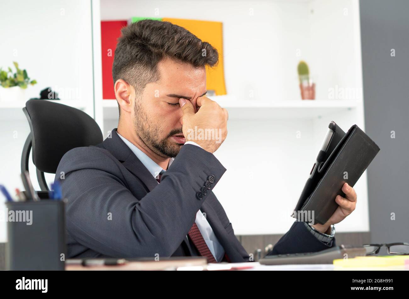 Portrait de l'homme fatigué travaillant avec la tablette numérique heures supplémentaires seul dans le bureau, en fermant les yeux et en pinçant son nez essayant de soulager le stress. Banque D'Images