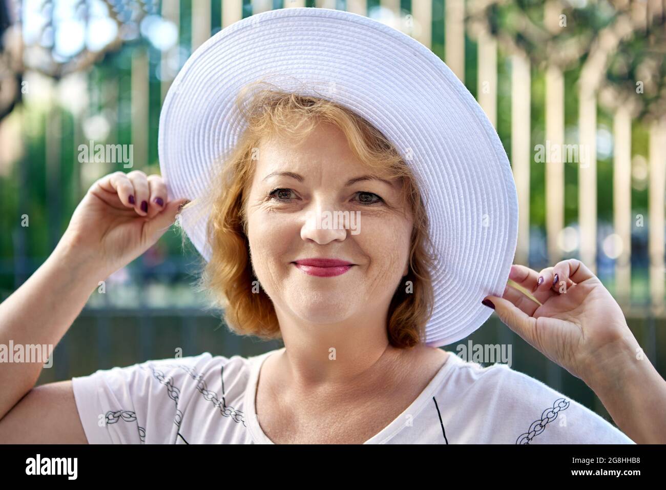 Belle femme âgée de 54 ans en chapeau blanc sourit tout en marchant dehors. Banque D'Images