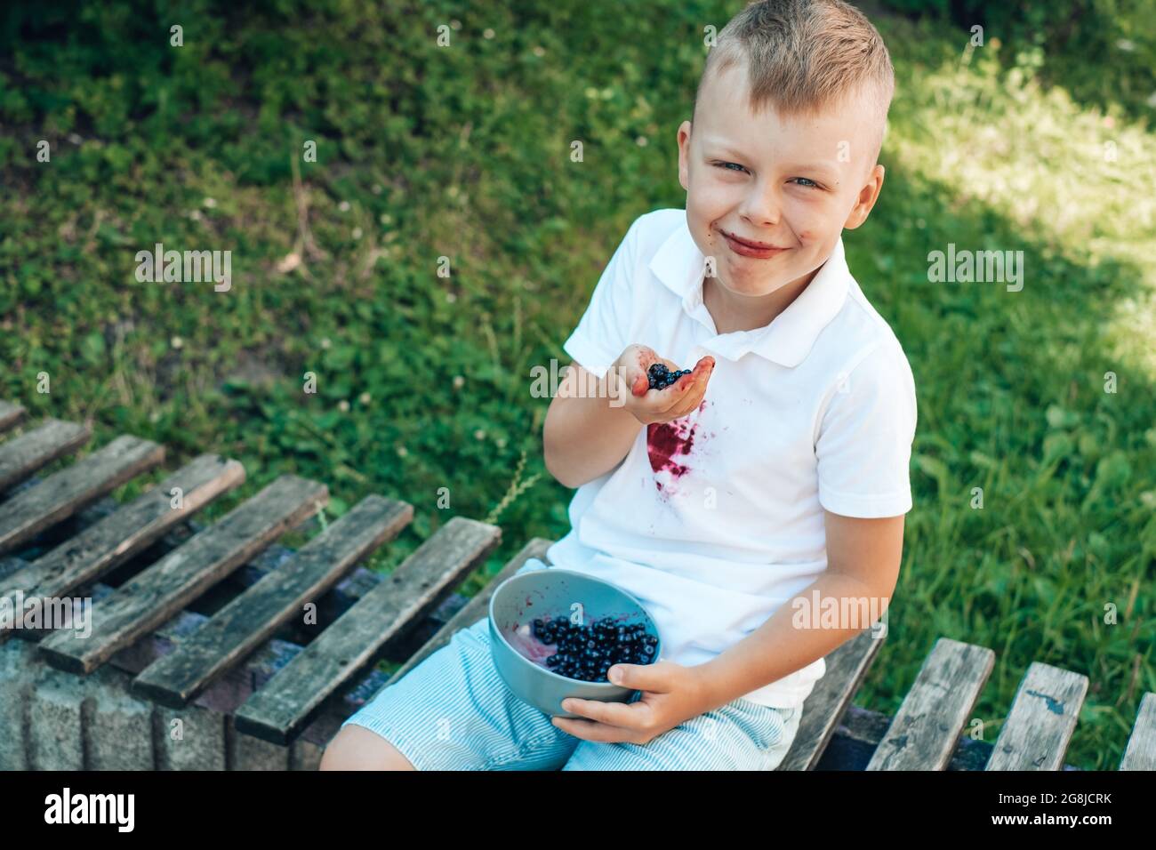 Joli garçon mangeant des baies fraîches avec ses mains et assis sur un banc en bois. À l'extérieur. Concept de nettoyage des taches Banque D'Images