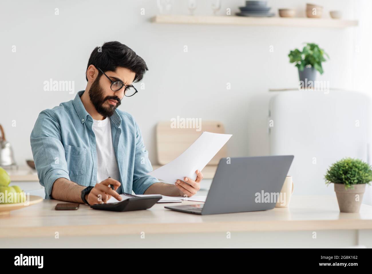 Payer des factures, des taxes à la maison en ligne. Jeune homme arabe utilisant une calculatrice et un ordinateur portable, assis à la cuisine, espace de copie Banque D'Images