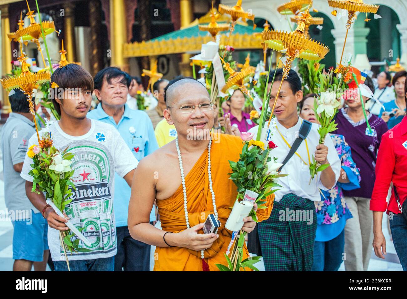 Jeune moine bouddhiste spectaculaire dans des robes orange avec des amis portant des offrandes de guirlande de fleurs et des parasols dorés, Pagode Shwedagon, Yangon, Myanmar Banque D'Images