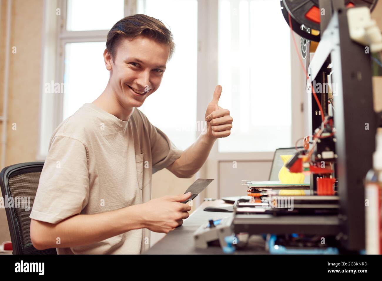 Jeune homme joyeux regardant la caméra avec le sourire et en faisant des gestes avec le pouce vers le haut tout en imprimant des modèles 3D à la maison Banque D'Images