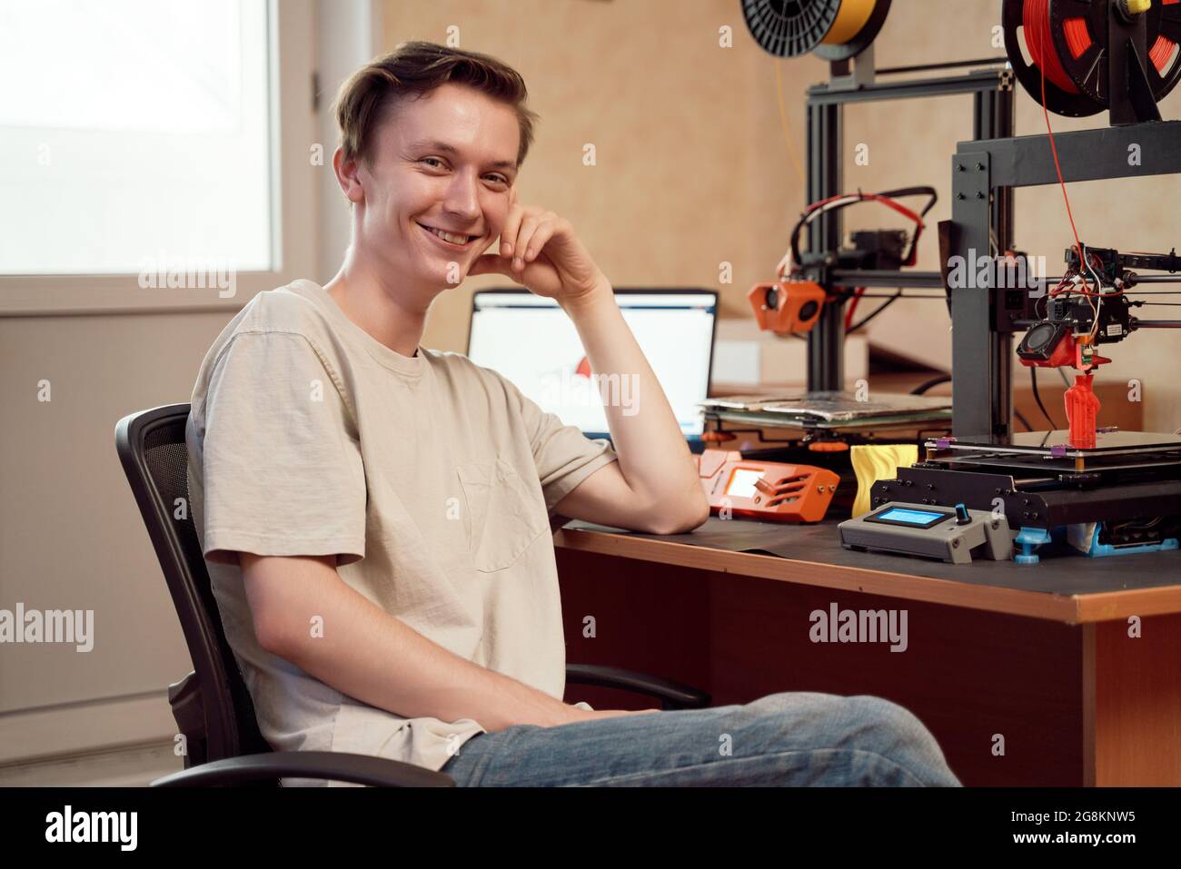 Un jeune homme heureux souriant et regardant l'appareil photo tout en étant assis à table avec des imprimantes 3D et un netbook à la maison Banque D'Images
