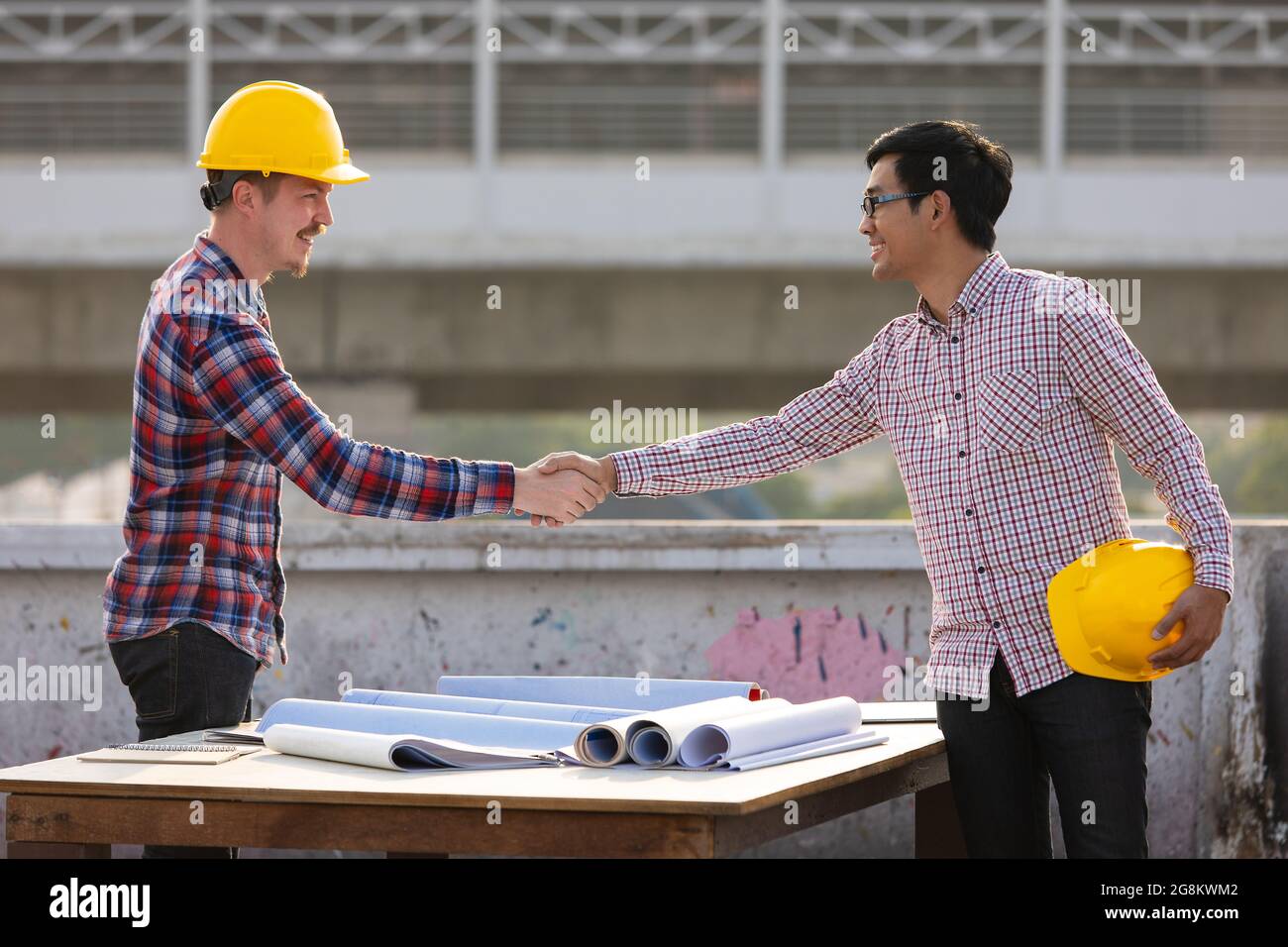 Deux ingénieurs, asiatiques et caucasiens se secouant la main après le travail est terminé sur le toit du chantier de construction avec de beaux rayons du soleil en arrière-plan. Banque D'Images