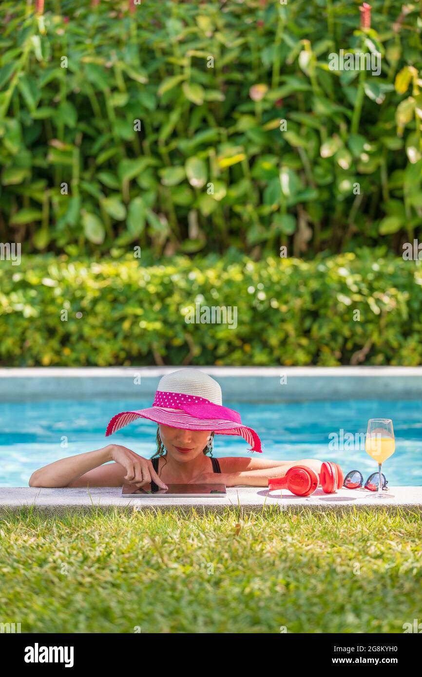 Moyenne d'âge 40 ans femme porter un grand chapeau et un bikini debout dans  la piscine à la lumière du soleil et en utilisant un ordinateur tablette.  Il y a un casque