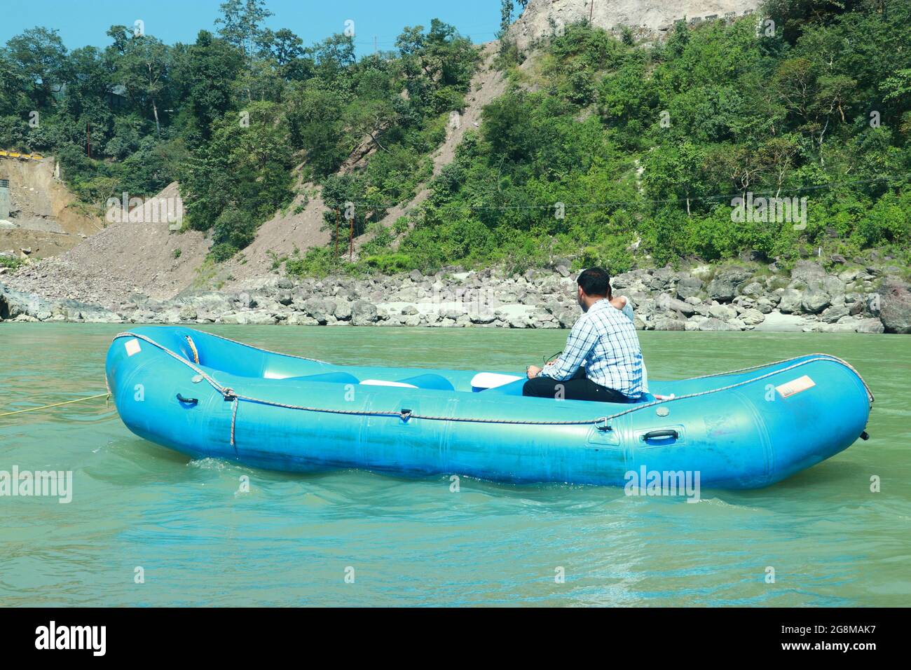 Jeune homme assis dans un bateau bleu et gonflable dans la rivière près de la terre et des arbres verts le jour d'été Banque D'Images