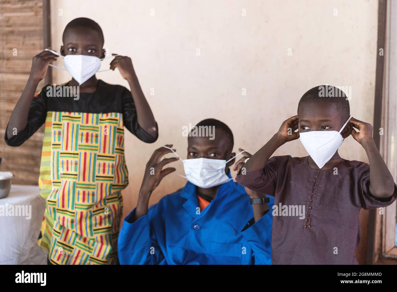 Dans cette image, un fournisseur de santé africain assis dans un manteau de laboratoire bleu montre comment porter et installer un masque antiviral de protection avec le soutien de deux Banque D'Images