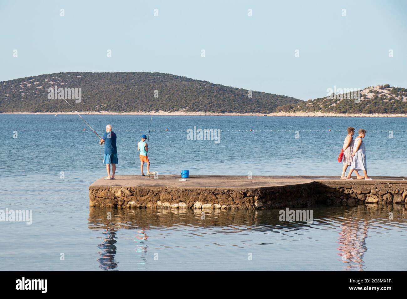 Vodice, Croatie - 8 juillet 2021 : homme âgé et jeune garçon pêchant sur une jetée tandis que les femmes âgées marchent Banque D'Images