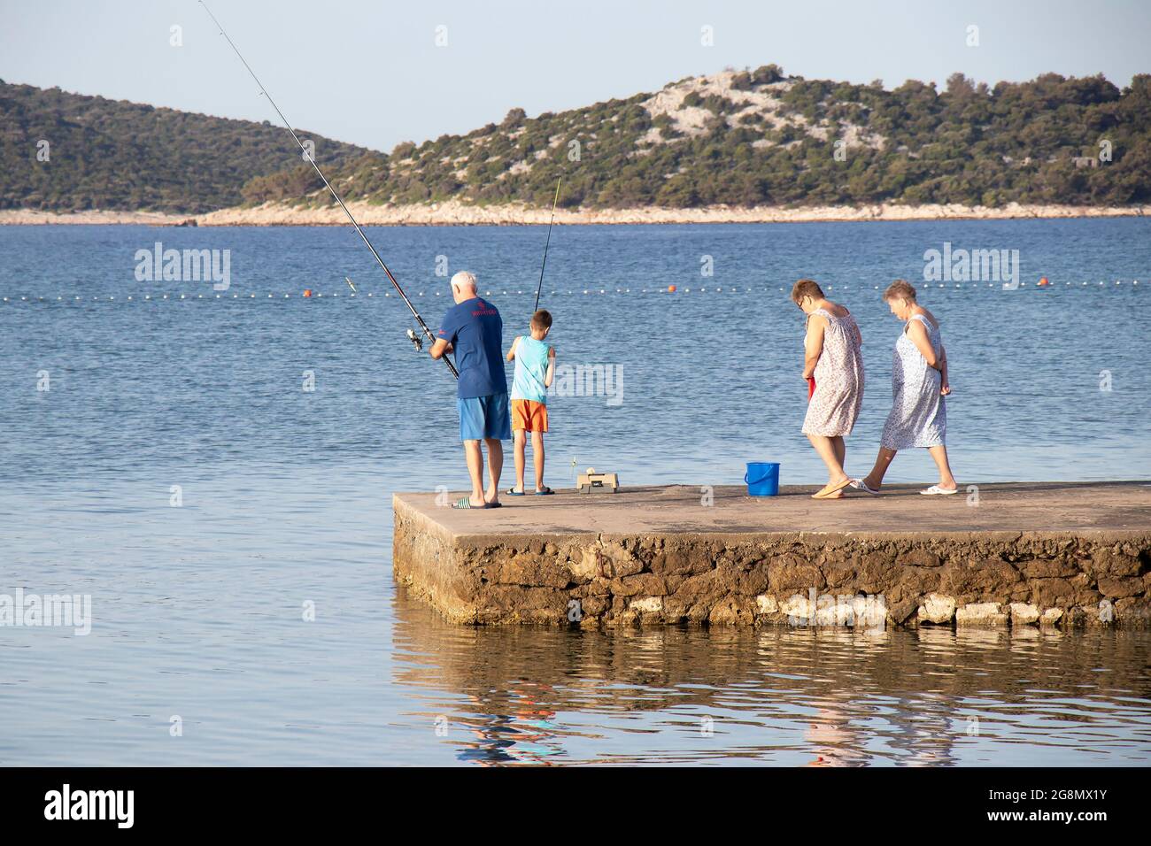 Vodice, Croatie - 8 juillet 2021 : homme âgé et jeune garçon pêchant sur une jetée tandis que les femmes âgées marchent Banque D'Images