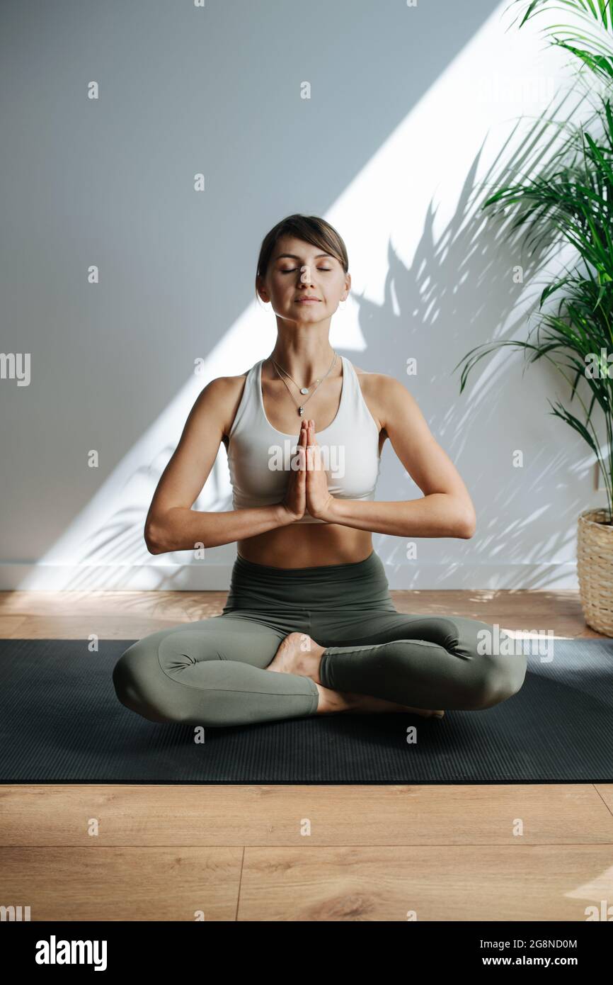 Calme femme de 30 ans pratiquant le yoga, méditant dans un grand studio. Vue frontale. Elle est assise en posture facile, ses yeux fermés, les mains jointes. Banque D'Images