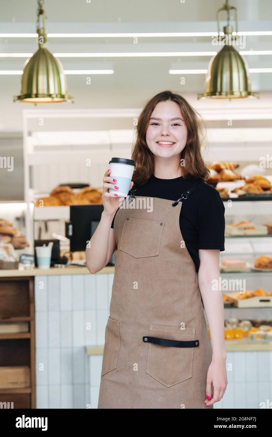 Jolie petite fille charmante posant avec une tasse de café en plastique à l'intérieur de son magasin. Elle porte un tablier et regarde la caméra. Sur les étagères, pleine de pâte. Banque D'Images