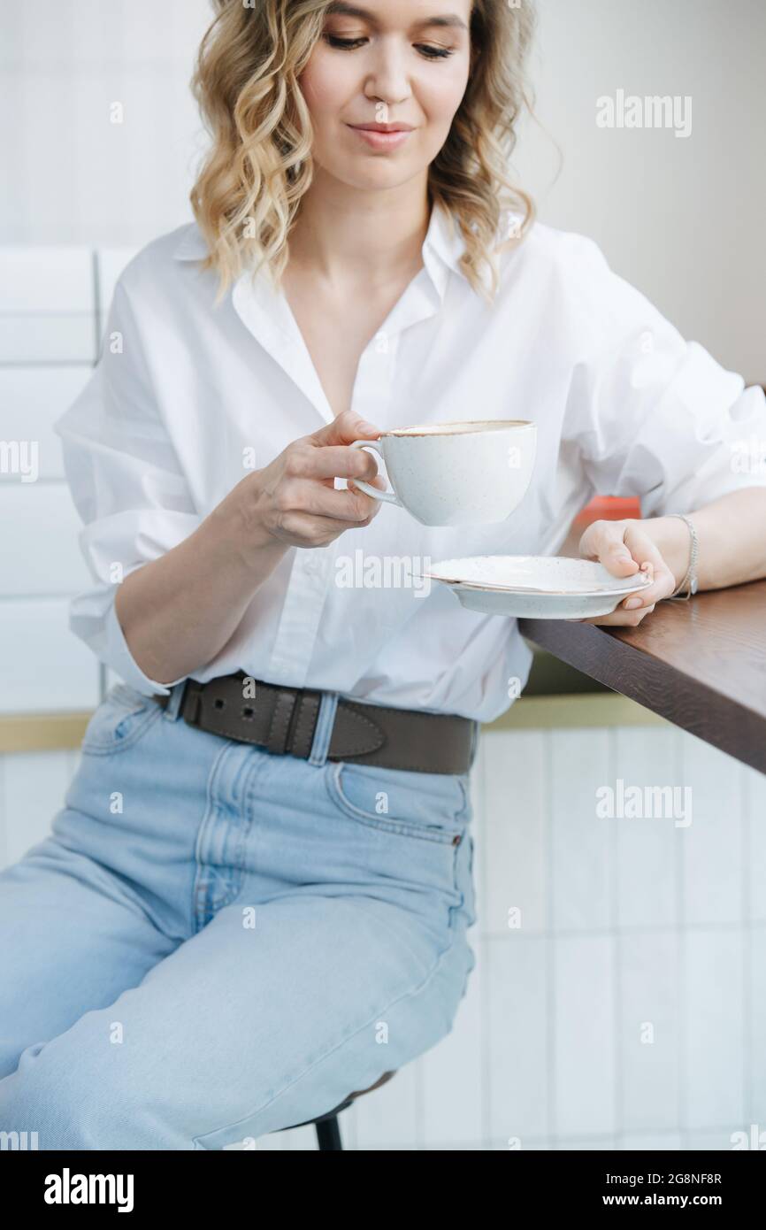 Femme solitaire blonde assise sur un haut tabouret dans un cafel, regardant une tasse de café. Elle porte une chemise bleue et un Jean léger. Banque D'Images