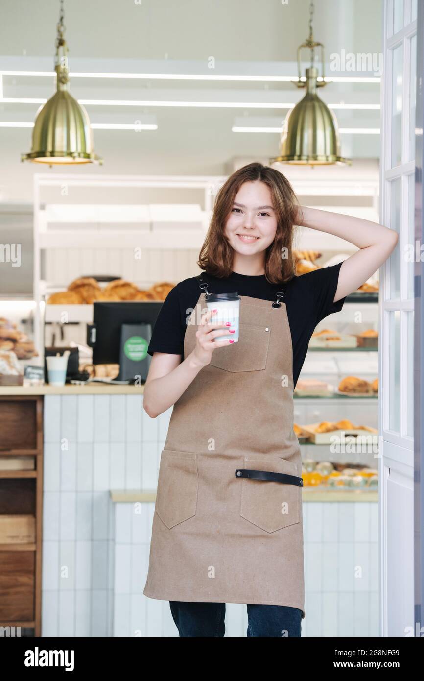 Belle jeune femme boulangère posant avec une tasse de café en plastique à l'intérieur de son magasin. Elle porte un tablier, regarde la caméra, s'appuie sur un cadre de porte. Ove Banque D'Images