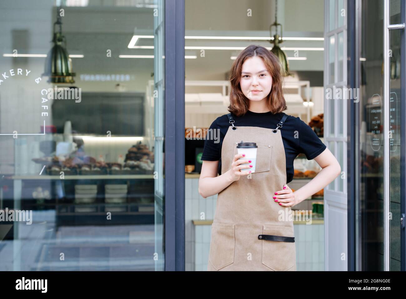 Jolie petite fille confiante posant avec une tasse de café en plastique devant son magasin. Elle porte un tablier et regarde la caméra. Banque D'Images