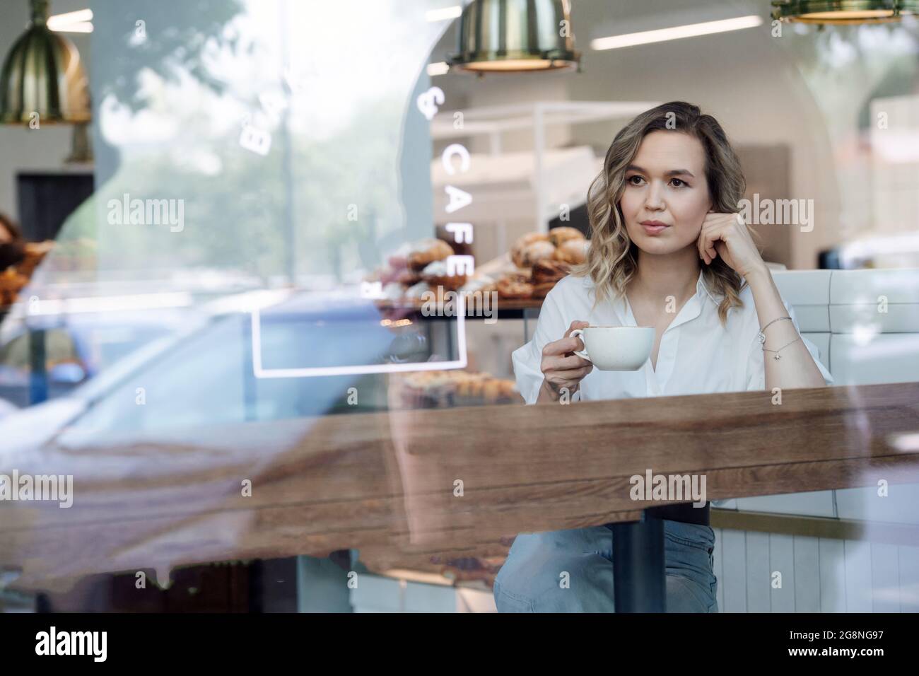 Paisible femme blonde appréciant son café dans un café, assis à côté d'une fenêtre, regardant à une distance. Elle porte une chemise bleue et un Jean léger. Banque D'Images